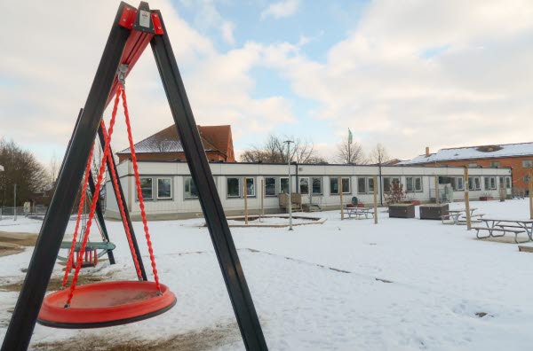 Röd gunga med paviljongbyggnad i bakgrunden. Foto.